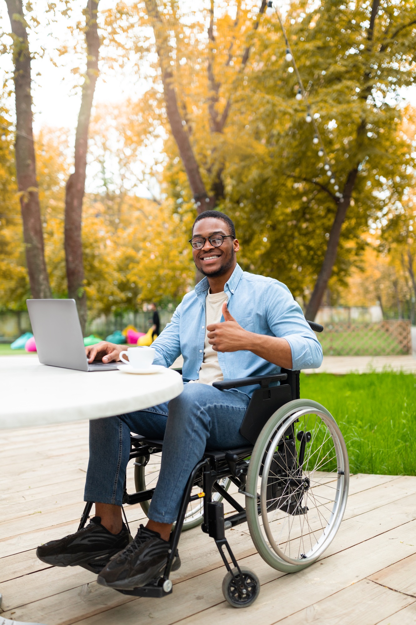 Online work for disabled people. Black guy in wheelchair working on laptop, showing thumb up at cafe